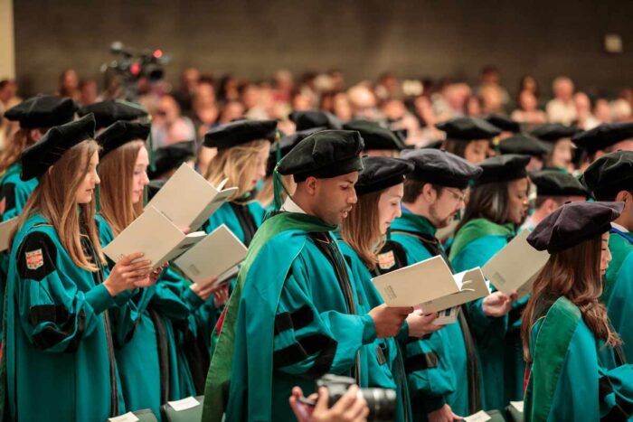 MD graduates in green regalia read from booklets.