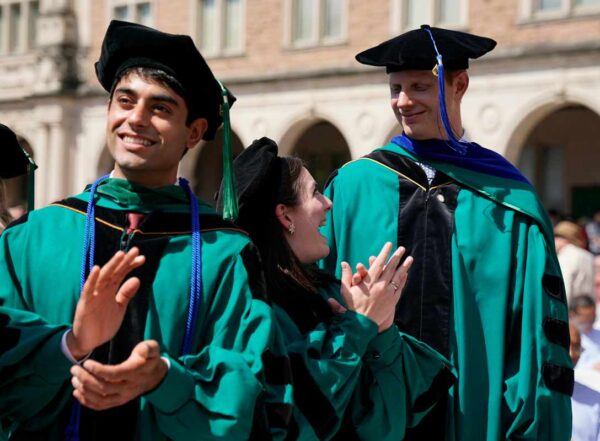 Three graduates in regalia stand and clap at outdoor ceremony