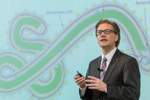 Dressed in a suit and tie, Lawrence presents in front of a green and blue slide depicting the Ebola virus