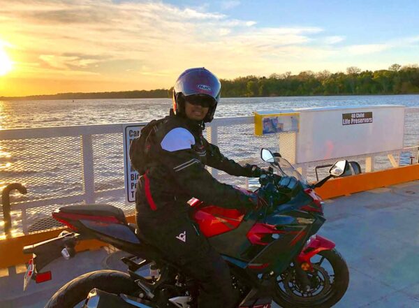 Guatam Adusumilli poses on his motorcycle wearing a jacket and helmet with a lake and sunset behind him