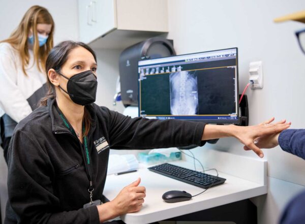 Devyani M. Hunt, MD, a professor of orthopedic surgery and medical director of the Living Well Center, talks with patient Zach Wood about pain issues. A computer monitor on the desk beside Dr. Hunt displays a spinal scan.