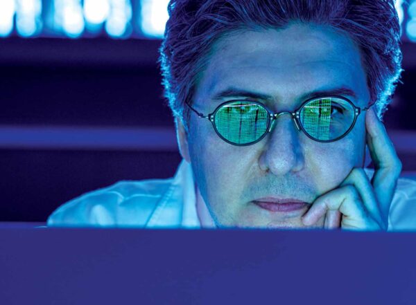 Viewed from over the top of a computer monitor, Ziyad Al-Aly, MD, is cast in indigo light. He looks intently into the monitor, facing the camera, cheek resting in his left hand. Rows of phosphor green numerals glow on his eyeglasses, reflected back from the screen before him.