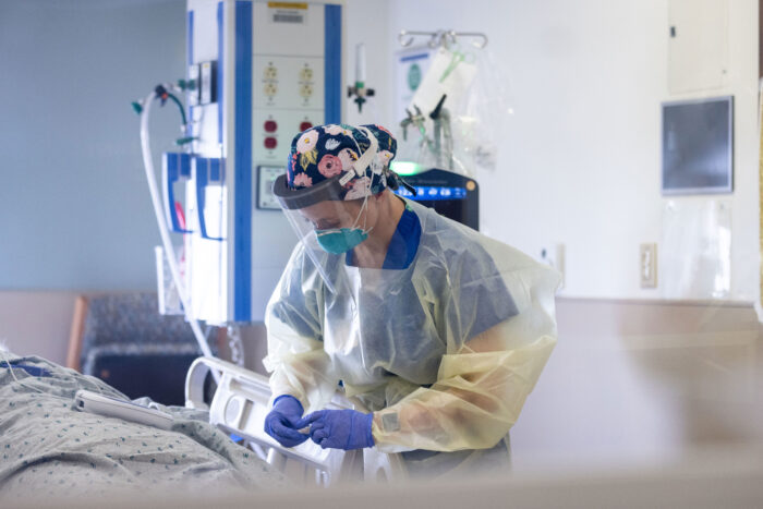 Nurse wearing full personal protective equipment bends over a patient bed in an ICU