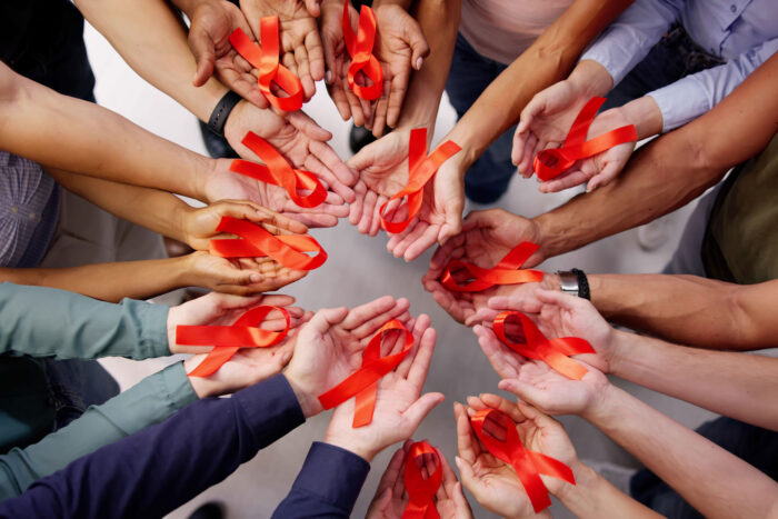 A circle of hands, all skin colors, holding red ribbons symbolizing HIV/AIDS