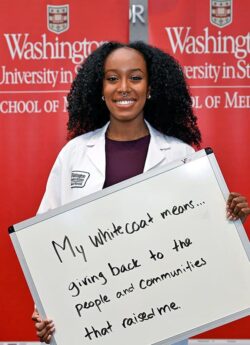 A smiling Black woman wearing a white physicians coat holds a handwritten sign that says: My white coat means giving back tot he people and communities that raised me.