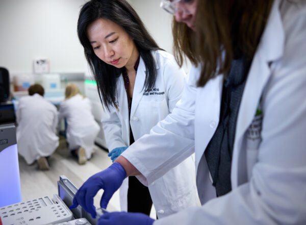 Lulu Sun, MD, PhD, and Shelly O’Laughlin, in the lab with white coats and blue gloves.