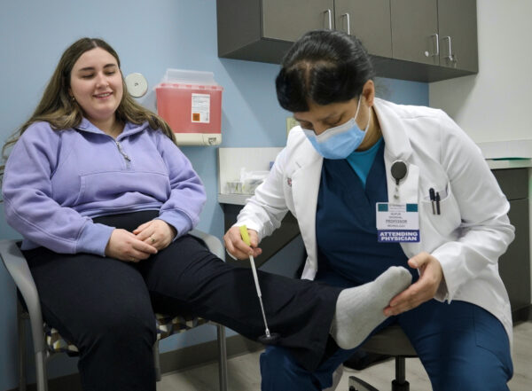 A patient sits with her leg outstretched while a doctor taps on her lower leg with an instrument.