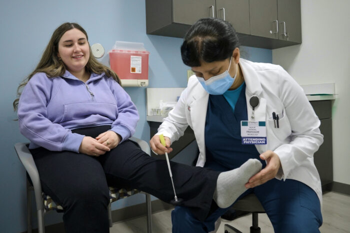 A patient sits with her leg outstretched while a doctor taps on her lower leg with an instrument.