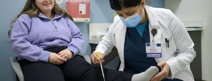 A patient sits with her leg outstretched while a doctor taps on her lower leg with an instrument.