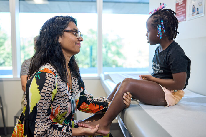 Female pediatric neurologist examines leg of a young girl in an exam room to assess movement.