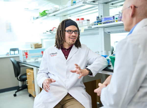 Two male scientists in white coats in a lab speaking.