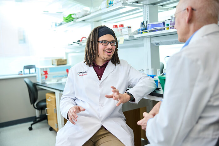 Two male scientists in white coats in a lab speaking.