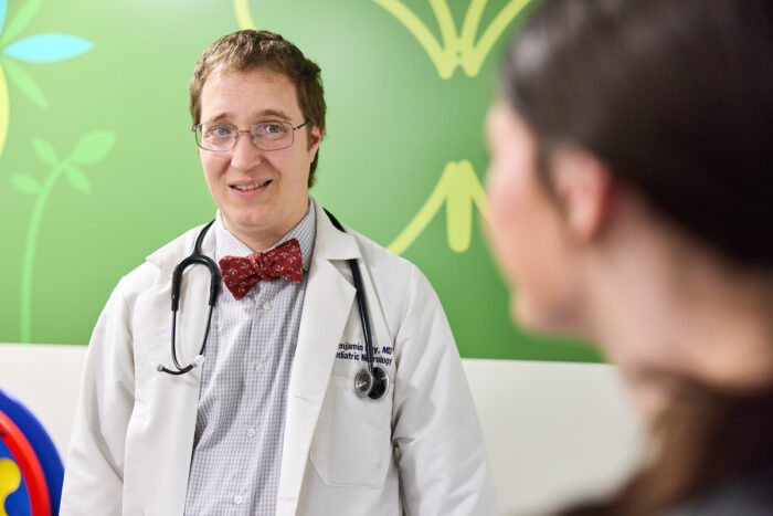 Benjamin Kay, MD, PhD, in white coat and bow tie with stethoscope around his neck, facing a pediatric patient