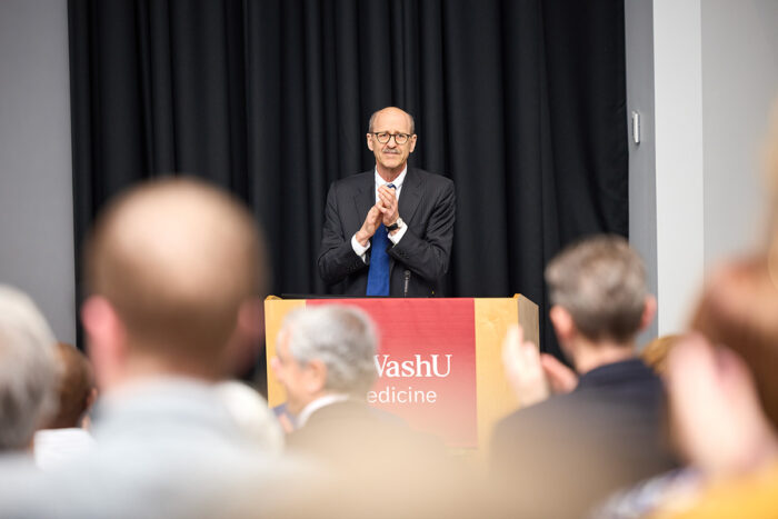 Dean in suit behind WashU Medicine lectern, facing crowd giving standing ovation