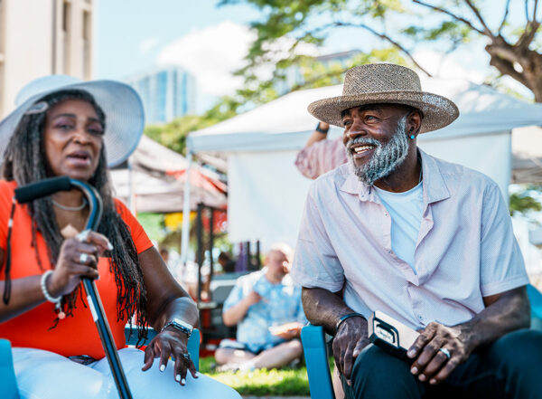 Smiling senior man sits at farmers market beside older woman in brightly colored shirt