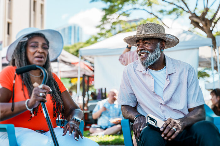 Smiling senior man sits at farmers market beside older woman in brightly colored shirt