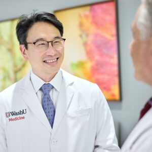 A man with dark hair and glasses, wearing a WashU Medicine white coat, smiles.