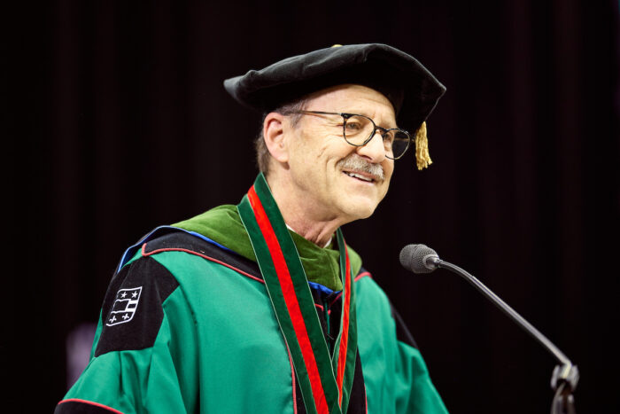 Dean David Perlmutter, dressed in Commencement regalia, speaks into a microphone.
