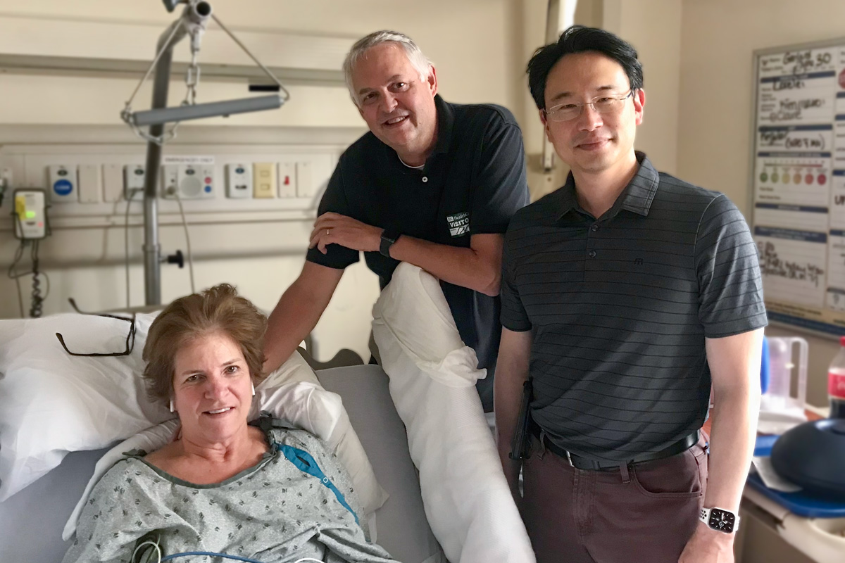 Patient in bed, spouse and surgeon posing bedside.