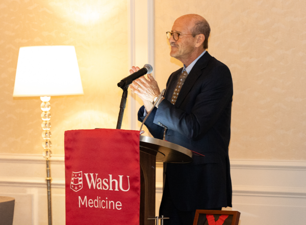 Man in dark suit and wearing glasses stands behind a podium, clapping