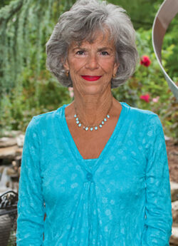 Woman standing in front of a garden, smiling. She is wearing a blue top.