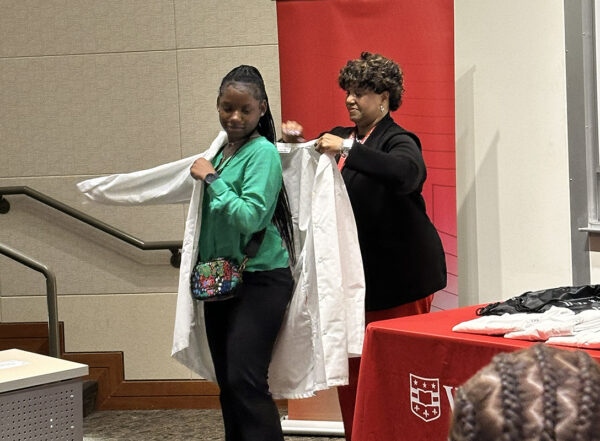 Woman standing behind high school girl, helping her into a medical white coat
