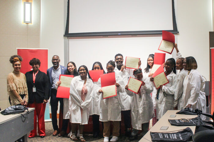 Three leaders stand beside ten high school students who are wearing their new medical white coats and waving their graduation certificates in the air.