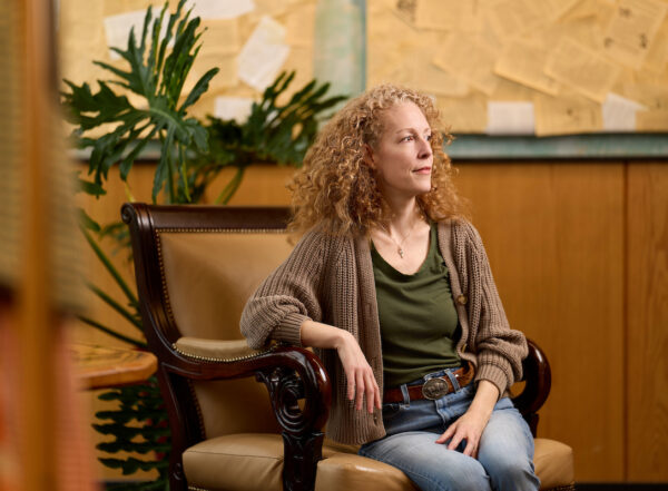 A person with curly hair sits in an ornate chair, gazing thoughtfully to the side in a warmly lit room with a large plant and a bulletin board in the background.