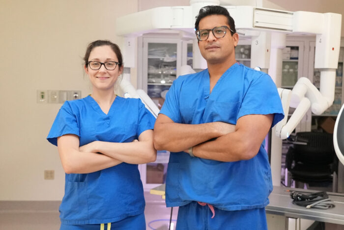 Female and male doctor in surgical scrubs pose with their arms crossed in an operating room.
