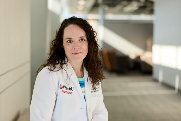 A person with curly hair wearing a white lab coat with "WashU Medicine" embroidered on it stands in a modern hallway.