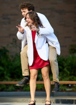 Two individuals in white lab coats, one carrying the other on their back, laugh and gesture joyfully outdoors.